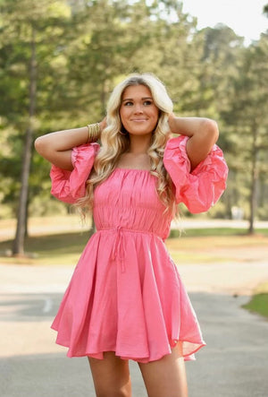 Puff Sleeves mini dress with tie at the waist. The neck line is elastic to provide for on the shoulder or off the shoulder wear. The hem line is not straight but is higher on the sides. The dress comes in a peachy pink color.